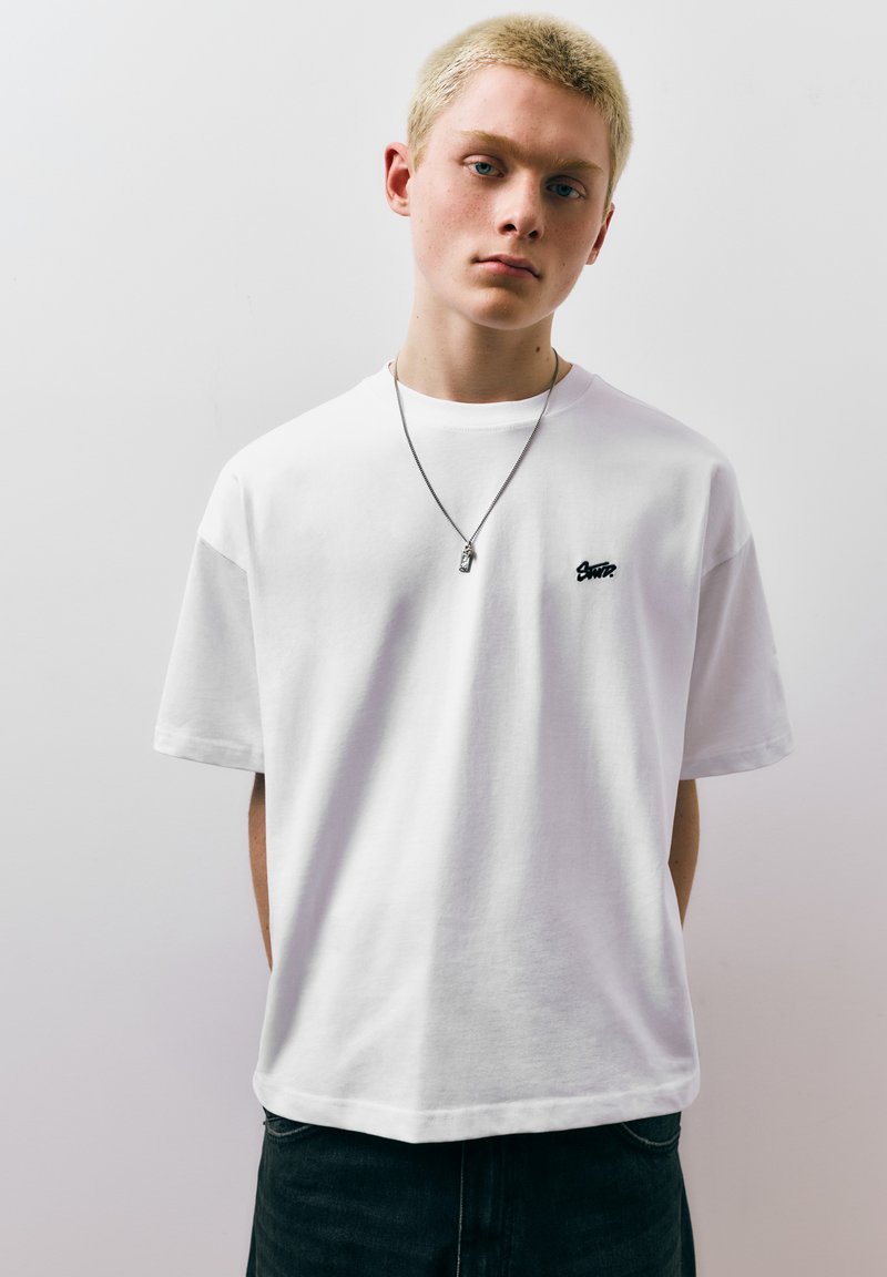 Jeune personne aux cheveux blonds courts portant un t-shirt blanc uni avec un petit logo noir et un collier en argent, debout devant un fond clair.