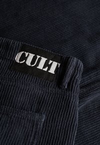 Granatowe spodnie sztruksowe o teksturowanej powierzchni, z czarną metką z napisem "CULT" na biało. Zawierają małą kieszonkę z przodu.