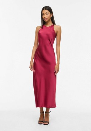 Kvinde står iført en ærmeløs, ankellang, silkeagtig mørkerød kjole med sorte hæle med ankelremme mod en ensartet hvid baggrund.