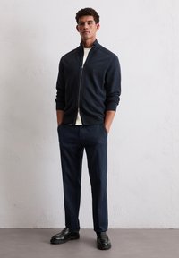 Marineblaues Zip-Up-Jacket mit gerippten Bündchen und Kragen, kombiniert mit marineblauen Hosen und schwarzen Schuhen. Glattes Material mit strukturierter Passform.