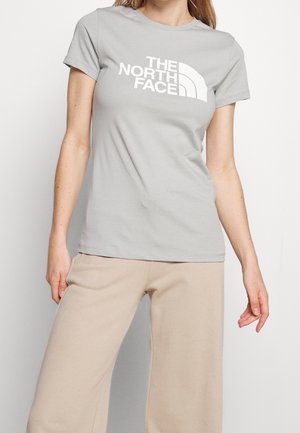 Kobieta ubrana w jasnoszarą koszulkę z krótkim rękawem z białym logo "THE NORTH FACE" oraz beżowe spodnie o szerokich nogawkach, stojąca na białym tle.