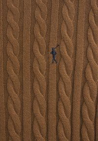 Suéter de punto trenzado en lana marrón, con un patrón texturizado de cables retorcidos y un logotipo de figura azul bordado en el frente.