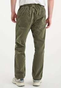 Man in olijfgroene elastische taillebroek en witte sneakers, gezien van achteren tegen een effen lichte achtergrond.