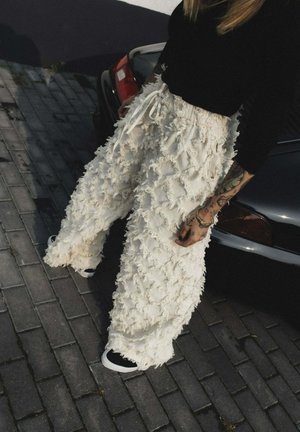 Osoba nosząca teksturowane, luźne białe spodnie i czarną bluzkę z długim rękawem stoi na szarym brukowanym chodniku obok czarnego samochodu.