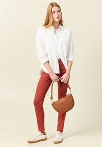 Borsa in pelle marrone con un design curvo, indossata con una camicia bianca e pantaloni color ruggine. Le sneakers presentano una combinazione di colori bianco e beige.