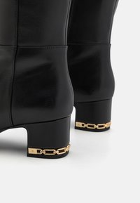 Botas de tobillo de cuero negro con tacón cuadrado, que presentan un acento de cadena dorada en la base. Textura suave con costuras cosidas.