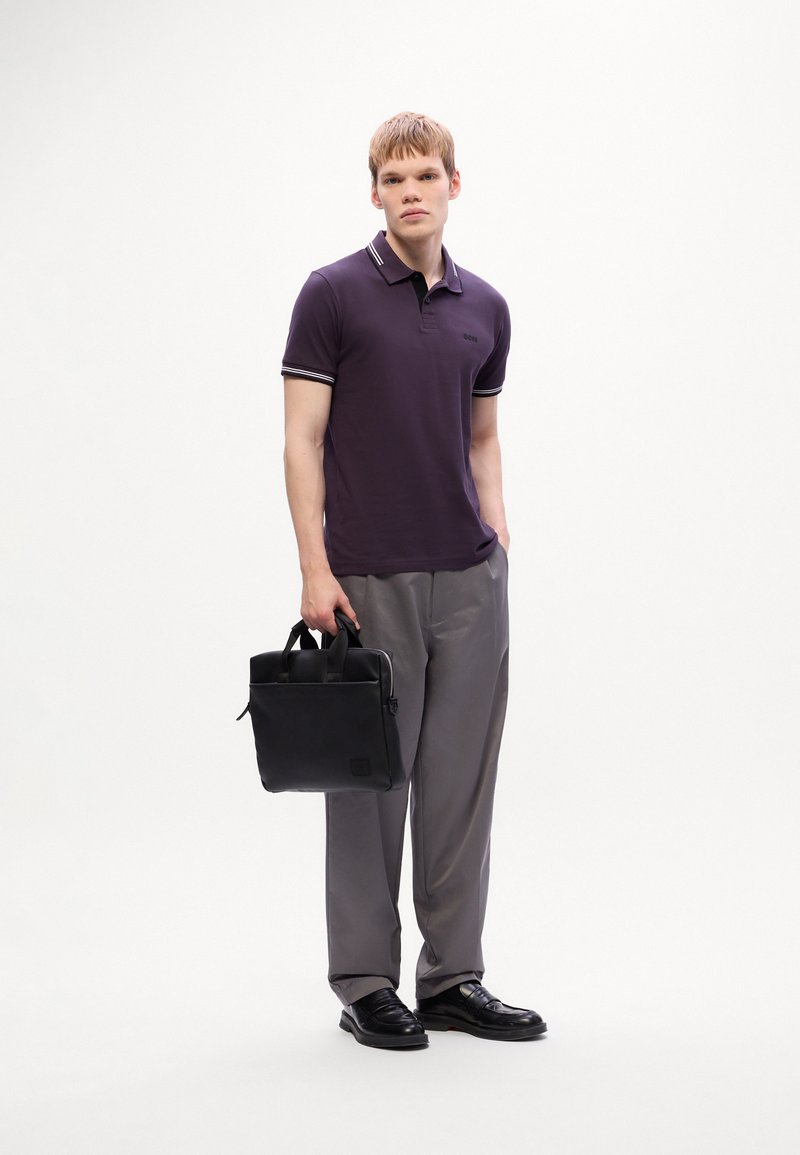 Jeune homme portant un polo violet et un pantalon gris, tenant une mallette noire dans une main, se tenant devant un fond blanc uni.