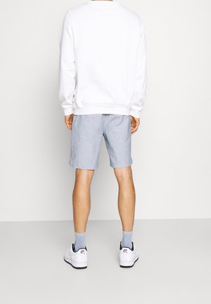 Muž v bílé mikině, světle modrých kraťasech, šedých ponožkách a bílých teniskách Nike Air, stojící zády k divákovi na jednobarevném pozadí.