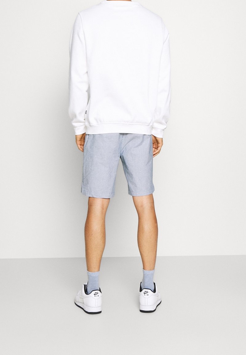 Mand iført hvid sweatshirt, lyseblå shorts, grå sokker og hvide Nike Air sneakers, stående med ryggen mod en ensfarvet baggrund.