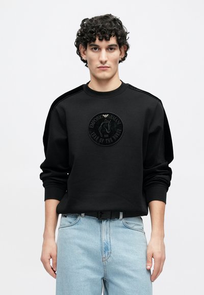 Melnā džemperī ar apaļu izšūtu logotipu ar zirgu un uzrakstu "Zirga gads". Mīksts audums un brīvs krūšu griezums, kombinēts ar gaiši zilām džinsa biksēm.