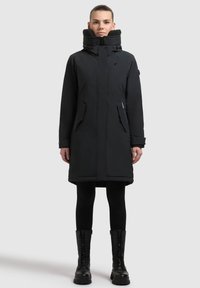 Parka negra con capucha, cierre de cremallera y a presión, cuello texturizado y bolsillos frontales. Lleva puesta sobre leggins negros y botas con cordones.