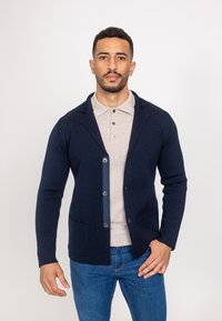 Tumesinine kudumiga kardigan, millel on pikad varrukad, šaalikaelus ja nööbiga kinniti. Omab tumedat sinist nööbiliistu ja kahte eesmist taskut. Kandes beeži polo.