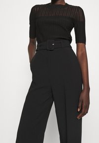 Top nero a costine con maniche corte, caratterizzato da righe verticali trasparenti. Pantaloni neri a vita alta con una cintura larga e tasche pronunciate.