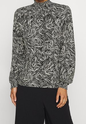 Femme portant un chemisier à manches longues avec un motif abstrait en spirale noir et blanc, des poignets froncés et un pantalon noir.