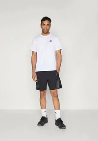 Tricou sportiv alb cu un logo negru, asortat cu pantaloni scurți negri cu dungi verticale, șosete albe și pantofi negri.