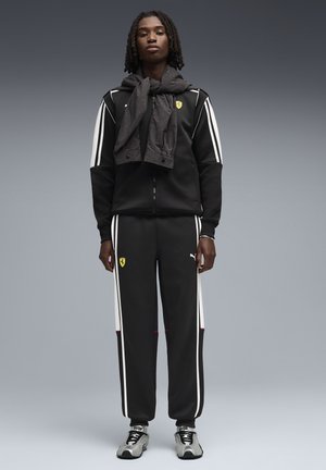 Musta verkkatakki valkoisilla raidoilla, Ferrari-logolla ja harmaa teksturoitu huivi kaulalla. Varustettu urheilukengillä.