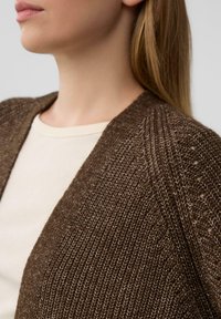 Brauner Strickcardigan mit offenem Vorderteil, strukturiertem Gewebe und subtiler Ausstrahlung; über einem hellbeigen Hemd getragen.