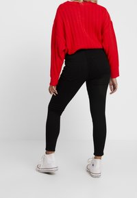 Jeans negros ajustados combinados con un suéter rojo de punto holgado. Los jeans tienen bolsillos traseros y una cintura alta, complementados con zapatillas blancas.