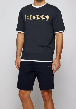 Camiseta de manga corta azul oscuro con un logo dorado de "BOSS", superpuesta sobre un cuello redondo blanco. Combinada con pantalones cortos náuticos a juego que presentan un logo dorado.