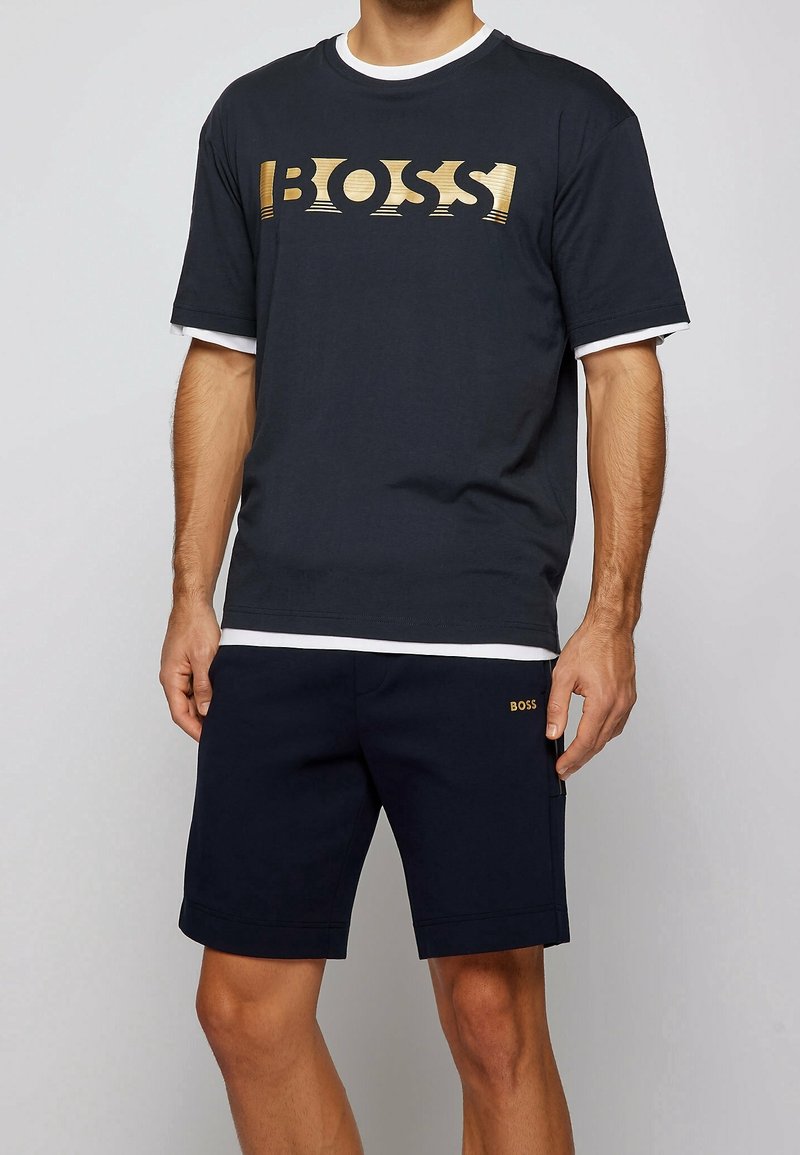Mörkblå kortärmad T-shirt med en guldig "BOSS"-logotyp, lagd över en vit rundad halsringning. Kombinerad med matchande marinblå shorts med en guldig logotyp.