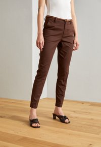 Pantalons marron sur mesure avec une coupe slim, ornés d'une ceinture à boucle circulaire, assortis avec des talons ouverts noirs et un top blanc ajusté.