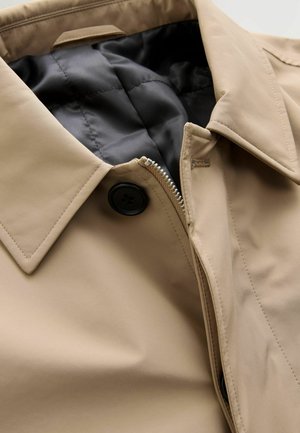 Beige Jacke mit Kragen, vorderem Reißverschluss und schwarzem Knopf. Verfügt über ein gestepptes schwarzes Innenfutter und eine glatte Außentextur.