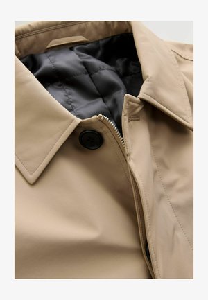 Beżowa kurtka z kołnierzem, przednim zamkiem błyskawicznym i czarnym guzikiem. Posiada pikowaną czarną podszewkę i gładką zewnętrzną fakturę.