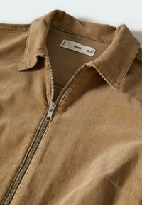 Chaqueta de pana marrón con cierre de cremallera, textura suave y cuello. Etiqueta visible en el interior del escote.