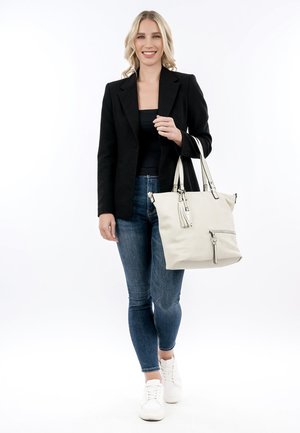 Blondīne sieviete melnā žaketē, melnā topā, zilās džinsās, baltās sporta kurpēs, tur lielu bēšu rokassomu, smaida, stāv uz balta fona.