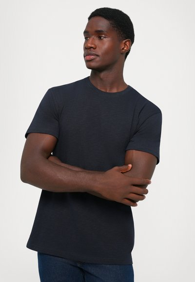 Man wearing a plain black short-sleeve t-shirt and blue jeans, standing with arms crossed, looking to the side against a plain background.