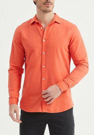 Homme boutonnant une chemise à manches longues orange vif avec col, portant un pantalon noir, sur un fond clair uni.