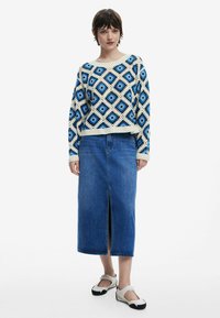 Pull en crochet avec un motif géométrique bleu et blanc, associé à une longue jupe en denim avec une fente devant. Des chaussures neutres complètent le look.