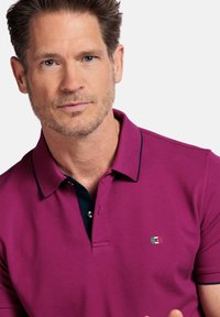Polo magenta avec un col bleu marine, arborant un petit logo brodé en rouge, vert et blanc. Tissu en coton, coupe classique.