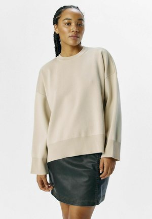 Beige oversized tröja med ribbade ärmslut och nederkant, kombinerad med en svart läderkjol som har en slät yta och en figursydd silhuett.
