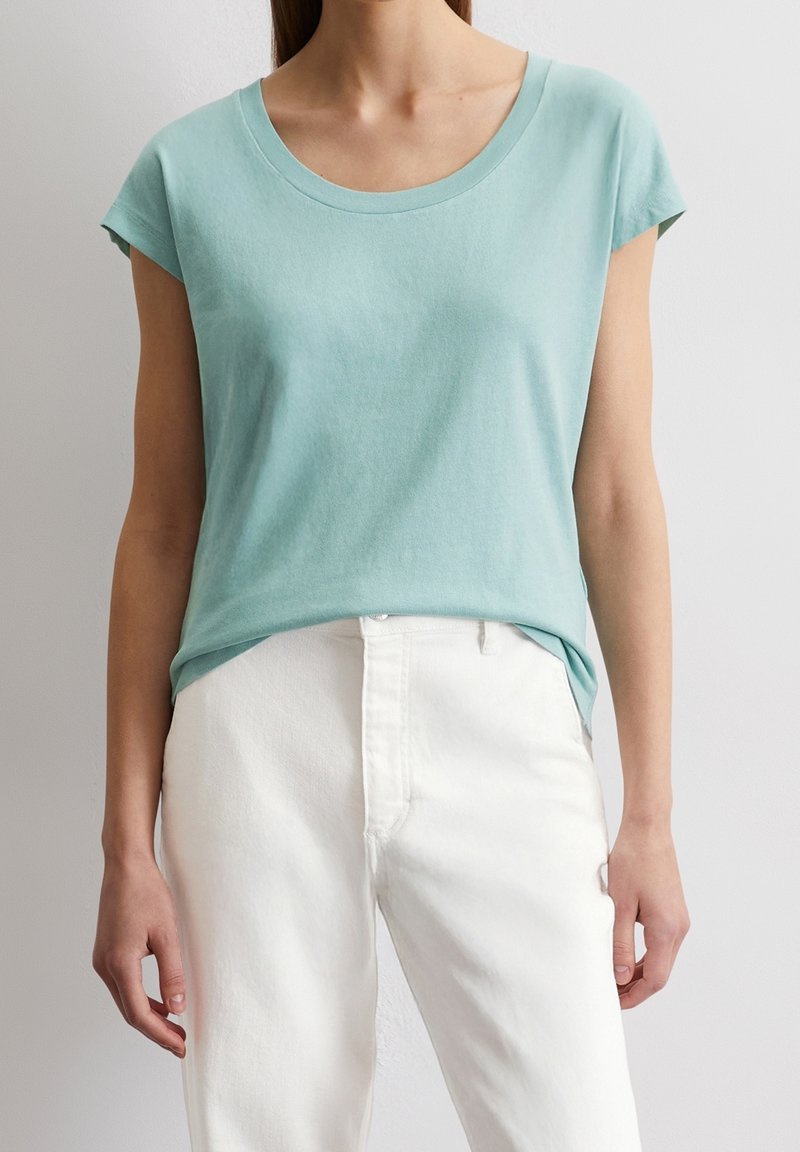 Ljusblå kortärmad T-shirt i mjuk bomull med rundad halsringning, kombinerad med vita byxor, visad mot en neutral bakgrund.