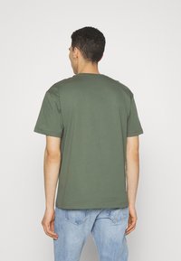 Oliwkowa bawełniana koszulka z okrągłym dekoltem, krótkimi rękawami i luźnym krojem. Noszona z jasnoniebieskimi dżinsami.