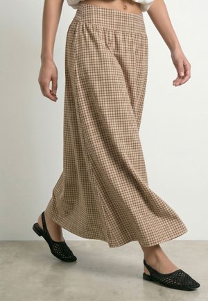 Personne portant un pantalon large à carreaux beige et marron et des ballerines slingback tissées noires marchant sur un sol lisse.