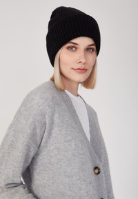 Berretto a coste nero indossato sopra capelli biondo chiaro, abbinato a un cardigan grigio con scollo a V e dettagli con bottoni.