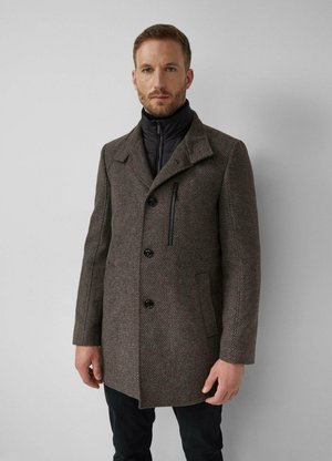 Man draagt een bruine wollen jas met textuur, een zwart ritsvak op de borst en een zwarte binnenjas, staande tegen een effen grijze muur.