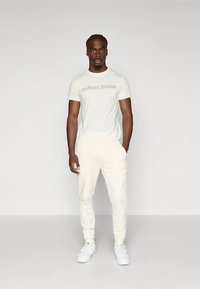 Svetlo bež majica s potiskom "BJÖRN BORG" na prsih, združena z ujemajočimi jogger hlačami. Gladek otip, preprost dizajn in beli supergi.