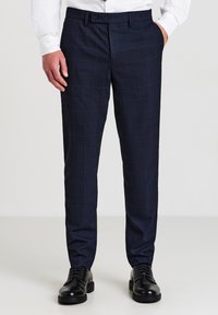 Mann in navyblauen karierten Anzughosen, weißem Hemd und schwarzen Anzugschuhen, der mit der rechten Hand in der Tasche vor einem weißen Hintergrund steht.