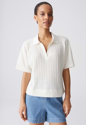 Frau trägt ein weißes kurzärmeliges, gestricktes Poloshirt mit Kragen und hellblaue Jeans-Shorts, steht vor einem einfarbigen Hintergrund.