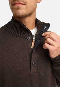 Bronze strikket sweater med høj ribbet krave, halvlange lynlås og trædetaljer. Har tre mørke knapper ved halsen.