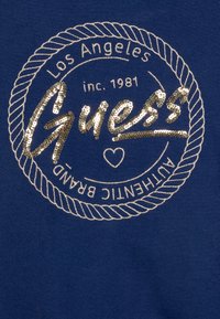 Navy blå sweatshirt med en rund logotyp med ett repdesign. "Guess"-texten är framträdande i guldpaljetter, med vita broderade detaljer.