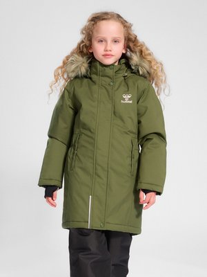 Kind met krullend haar, gekleed in een lange olijfgroene winterjas met bontkraag en een zwarte broek, staand voor een effen achtergrond.