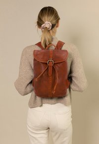 Sac à dos en cuir marron de forme arrondie, avec des bretelles réglables, un rabat de fermeture et un cordon de serrage. Finition texturée, accessoires en métal robustes.