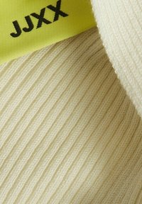 Krämfärgad ribbad tyg med en vikning i kanten och en gul etikett med fet svarta bokstäver "JJXX".