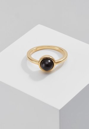 Bague en or avec une pierre ronde noire polie sertie dans un chaton simple, posée sur une surface géométrique blanche sur un fond gris.