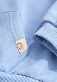 Vêtement tricoté bleu clair montrant un gros plan sur le poignet de la manche, le bord de la poche et une petite étiquette en tissu beige avec un logo partiel.