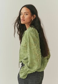 Jasnozielona, teksturowana bluzka z postrzępionymi detalami, długimi rękawami i marszczonym dołem z troczkiem. Noszona z ciemnymi spodniami w wzory.